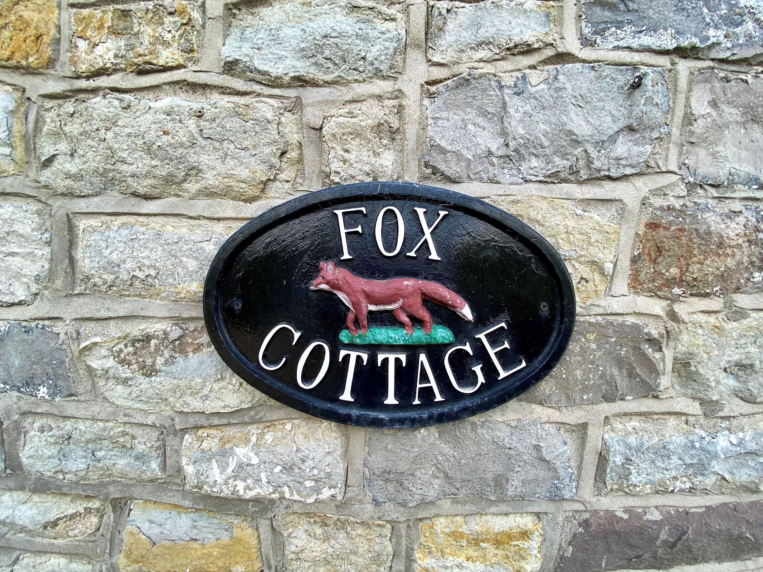 Segno in metallo nero raffigurante una volpe e riportante la scritta "FOX COTTAGE" fissato a un muro di pietra.