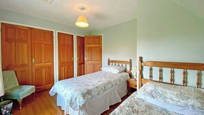 A bedroom with two single beds, a wooden wardrobe, a chair, and a bedside table with a lamp.