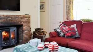 A cozy living room with a fireplace, a sofa with red cushions, a table with a floral tablecloth, and a flat-screen TV mounted on the wall. - Inverchroskie Cottage (Enochdhu, near Pitlochry)