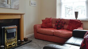 A living room with a fireplace, a red sofa, a black leather chair, and a window with blinds. - 3 bedroom accommodation in Keswick (Keswick)