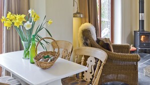 Une pièce avec une table blanche, des chaises en osier, un panier en osier, un vase avec des fleurs, une chaise en osier recouverte d’un plaid en fourrure, une lampe suspendue et un poêle à bois.