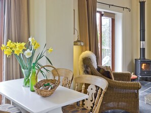 Ein Zimmer mit einem weißen Tisch, Korbst ühlen, einem Korb, einer Vase mit Blumen, einem Korbstuhl mit Felldecke, einer Hängelampe und einem Holzofen.