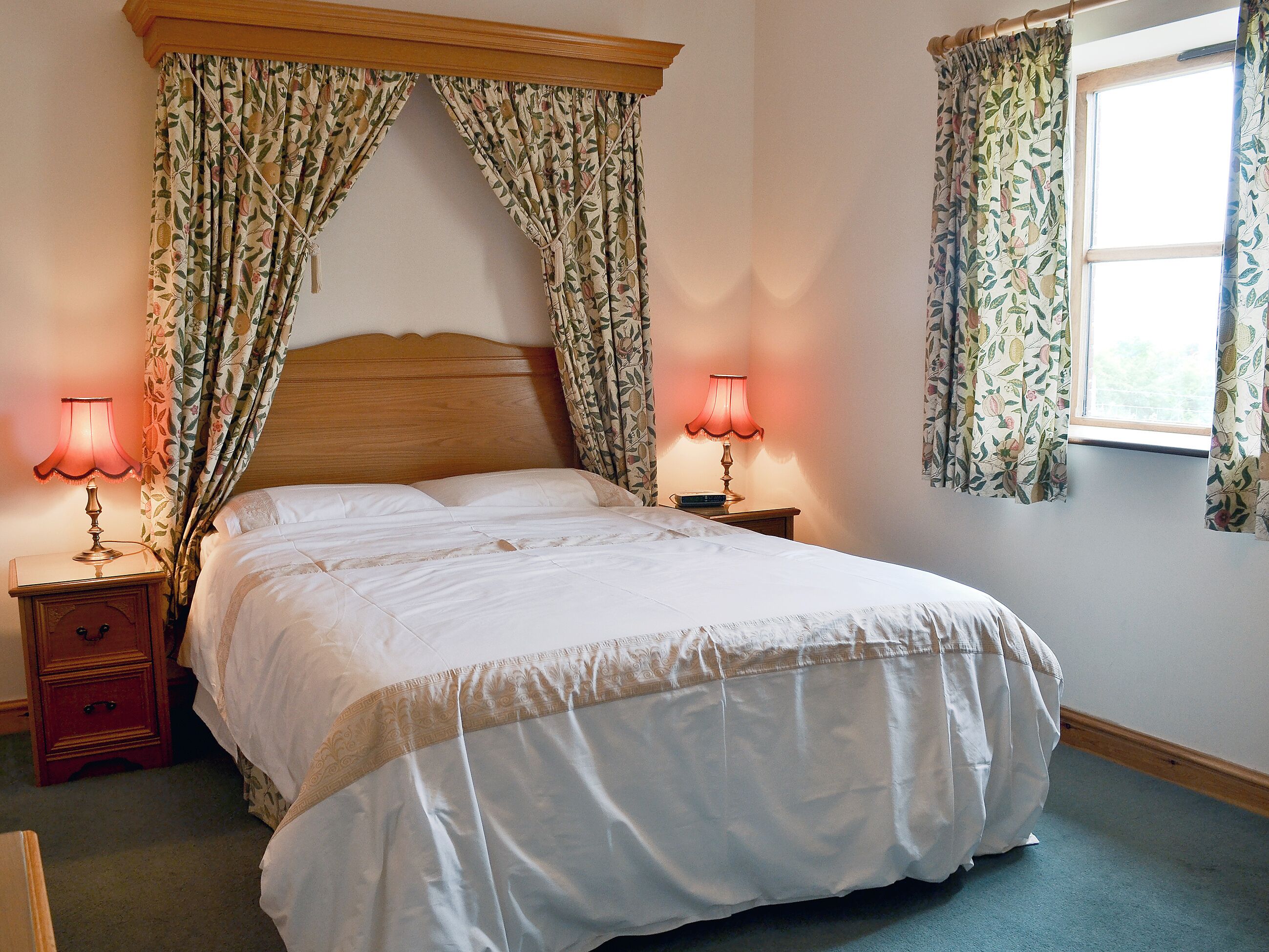 Ein Schlafzimmer mit einem großen Bett, zwei Nachttischen mit Lampen, floralen Vorhängen und einem Fenster mit Vorhängen.