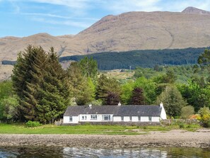 A white house with a sloped roof, situated by a body of water, surrounded by greenery and mountains in the background.