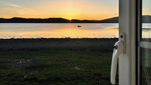 Vista de un lago al atardecer con un bote en el agua, una orilla cubierta de césped y una cadena montañosa al fondo.