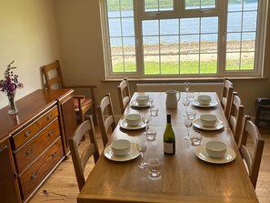 Une table à manger dressée pour six personnes, avec assiettes, verres et une bouteille, placée près d’une fenêtre donnant sur un lac.