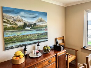 A dining area with a painting of a landscape, a wooden table with fruits, wine bottles, and a bowl of apples. - 3 bedroom accommodation in Taynuilt (Taynuilt)