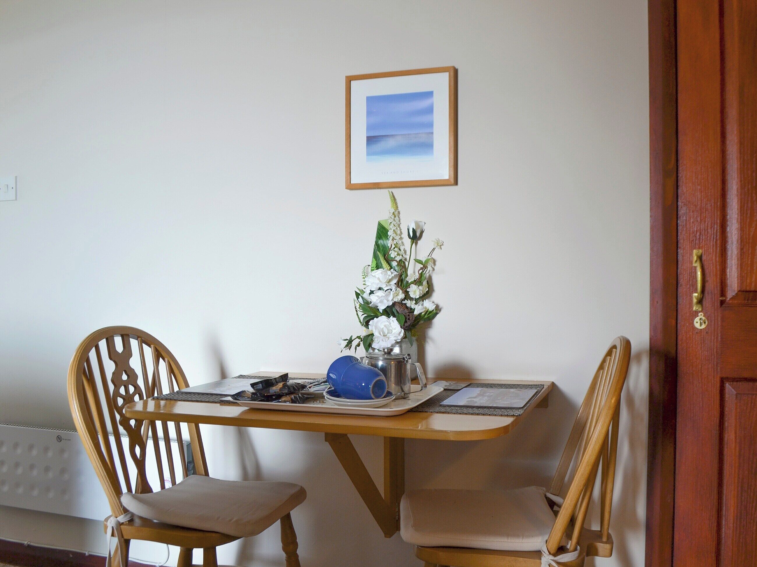 Un coin repas comprenant deux chaises en bois, une table ornée d’un vase de fleurs, un tableau encadré accroché au mur et une porte en bois.
