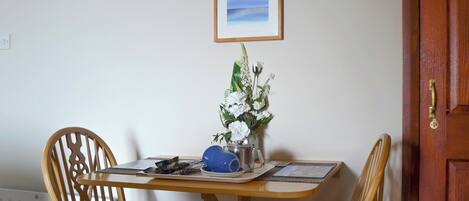 Un coin repas comprenant deux chaises en bois, une table ornée d’un vase de fleurs, un tableau encadré accroché au mur et une porte en bois.