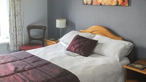 Una cama bien hecha con una colcha blanca y granate, un cabecero de madera y una sola almohada decorativa. Un mesita de noche con una lámpara y una pequeña mesa con un libro están al lado de la cama. Un cuadro floral cuelga en la pared sobre la cama.
