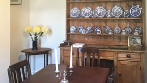 Un comedor con una mesa de madera, sillas, un jarrón con flores y una repisa con platos azules y blancos.