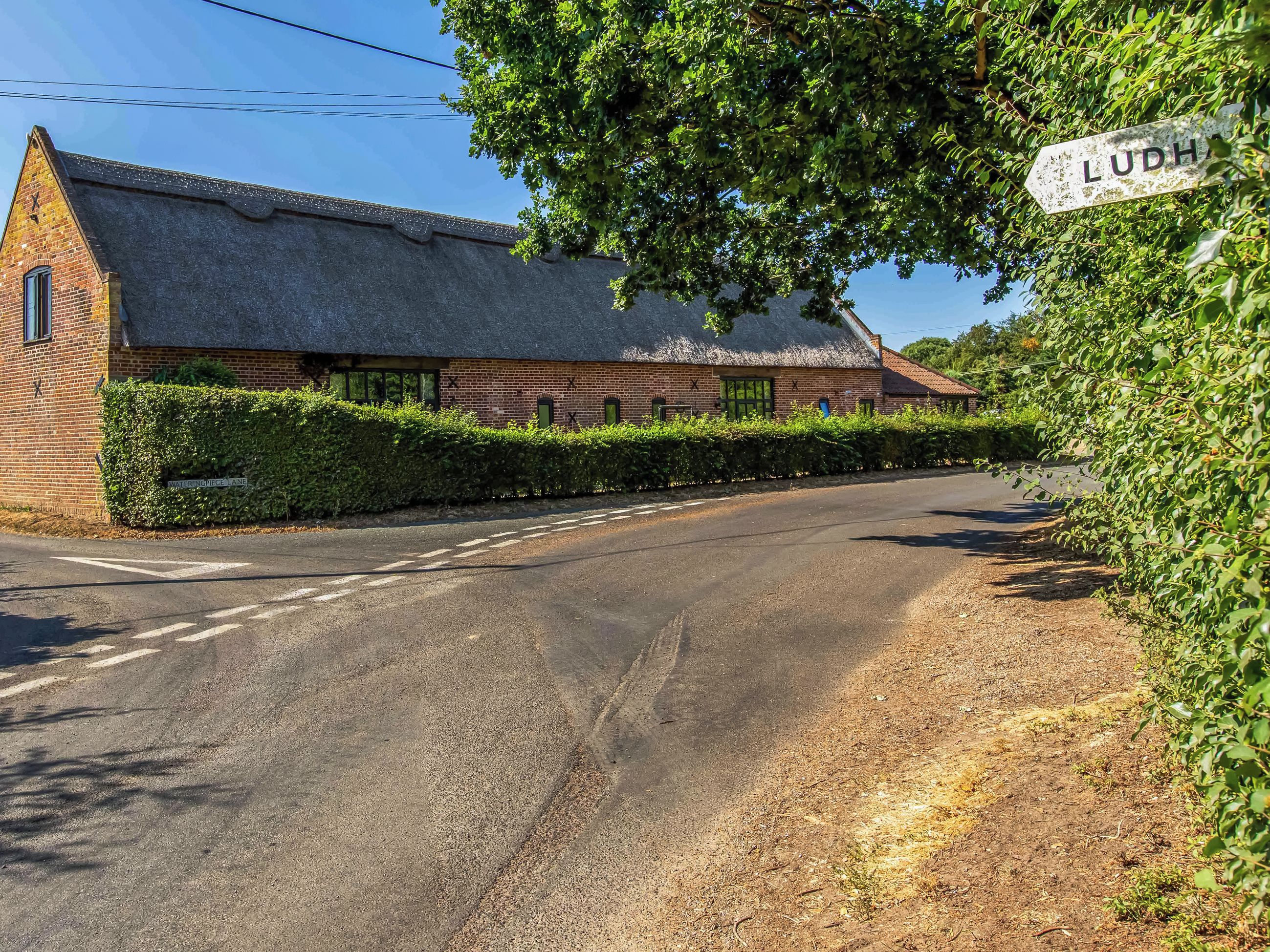A rural road leading to a brick building with a thatched roof, surrounded by greenery and a clear sky.