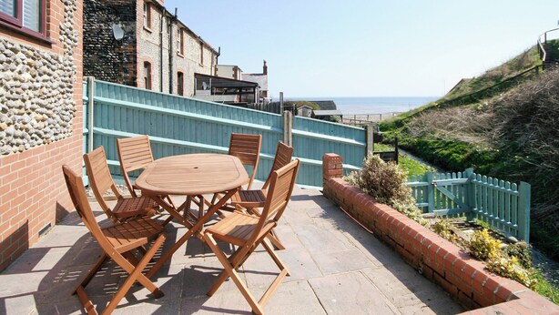 A patio with a round wooden table and chairs, overlooking a coastal area with a view of the sea and a grassy hill. - Beach Retreat - E5558 (Mundesley)