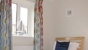 A bedroom with a bed, a window, curtains, and a small bedside table. - Beach Retreat - E5558 (Mundesley)
