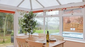 Un invernadero con una mesa de madera dispuesta para comer, amplios ventanales y vistas a un jardín y un muro de ladrillo.