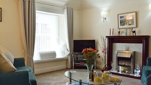 Un salon comprenant un canapé, une table basse avec des livres et un vase, un téléviseur à écran plat, une cheminée et une fenêtre avec des rideaux.