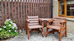A wooden outdoor seating area with two chairs and a table, a wooden fence, and a building with a window.