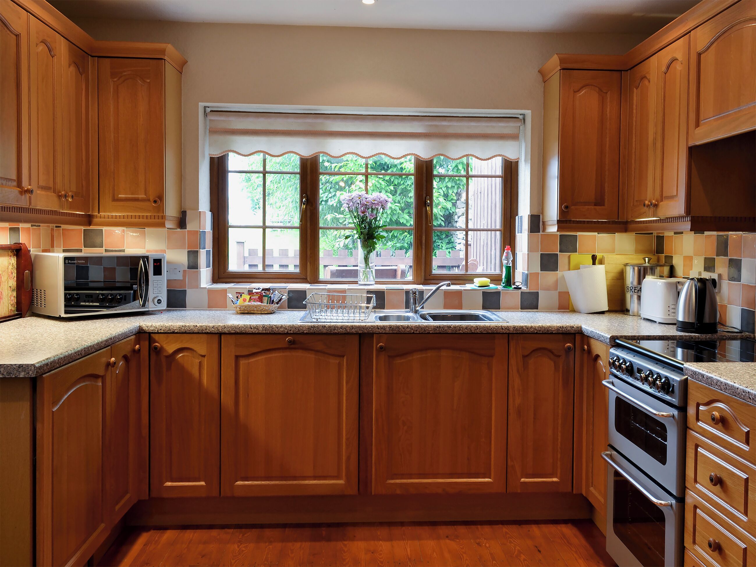 A kitchen with wooden cabinets, a microwave, a sink, and a window with a view of greenery.