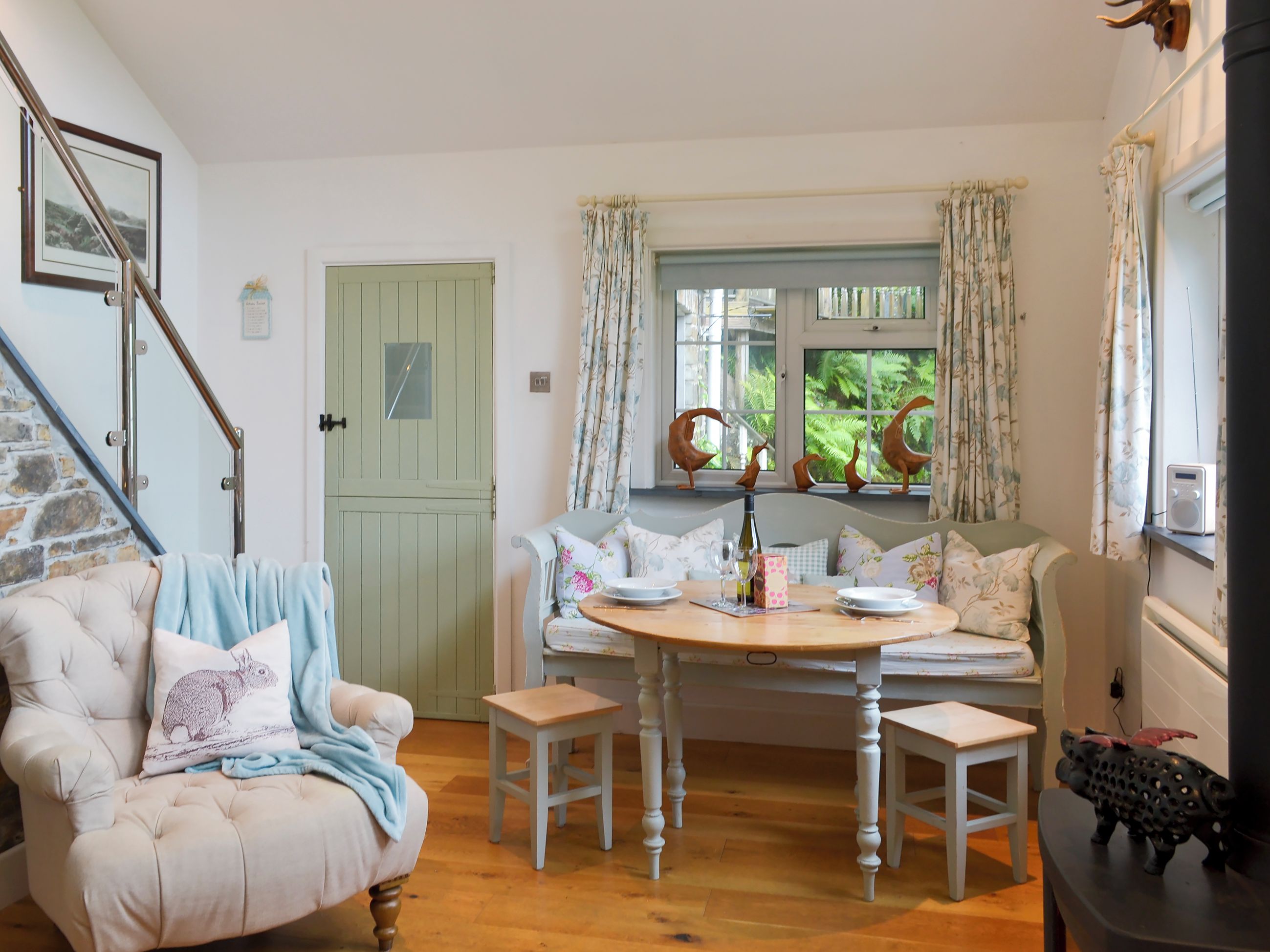 Une pièce chaleureuse avec une table à manger dressée pour deux personnes, un fauteuil moelleux, un petit tabouret en bois et une fenêtre avec des rideaux.
