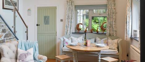 Une pièce chaleureuse avec une table à manger dressée pour deux personnes, un fauteuil moelleux, un petit tabouret en bois et une fenêtre avec des rideaux.