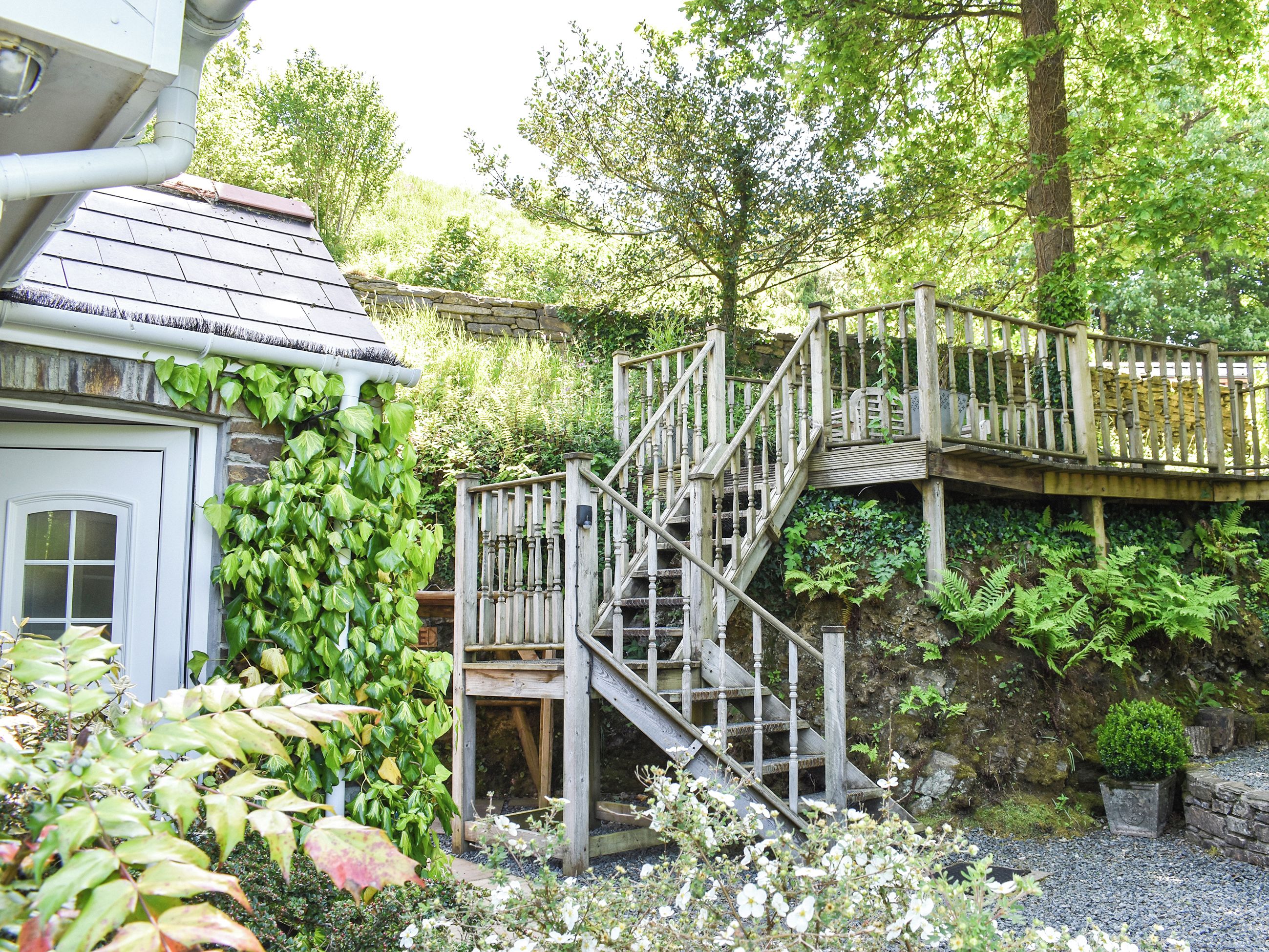 En trætrap fører op til en stenvæg med frodig vegetation og en bygning med en hvid dør.
