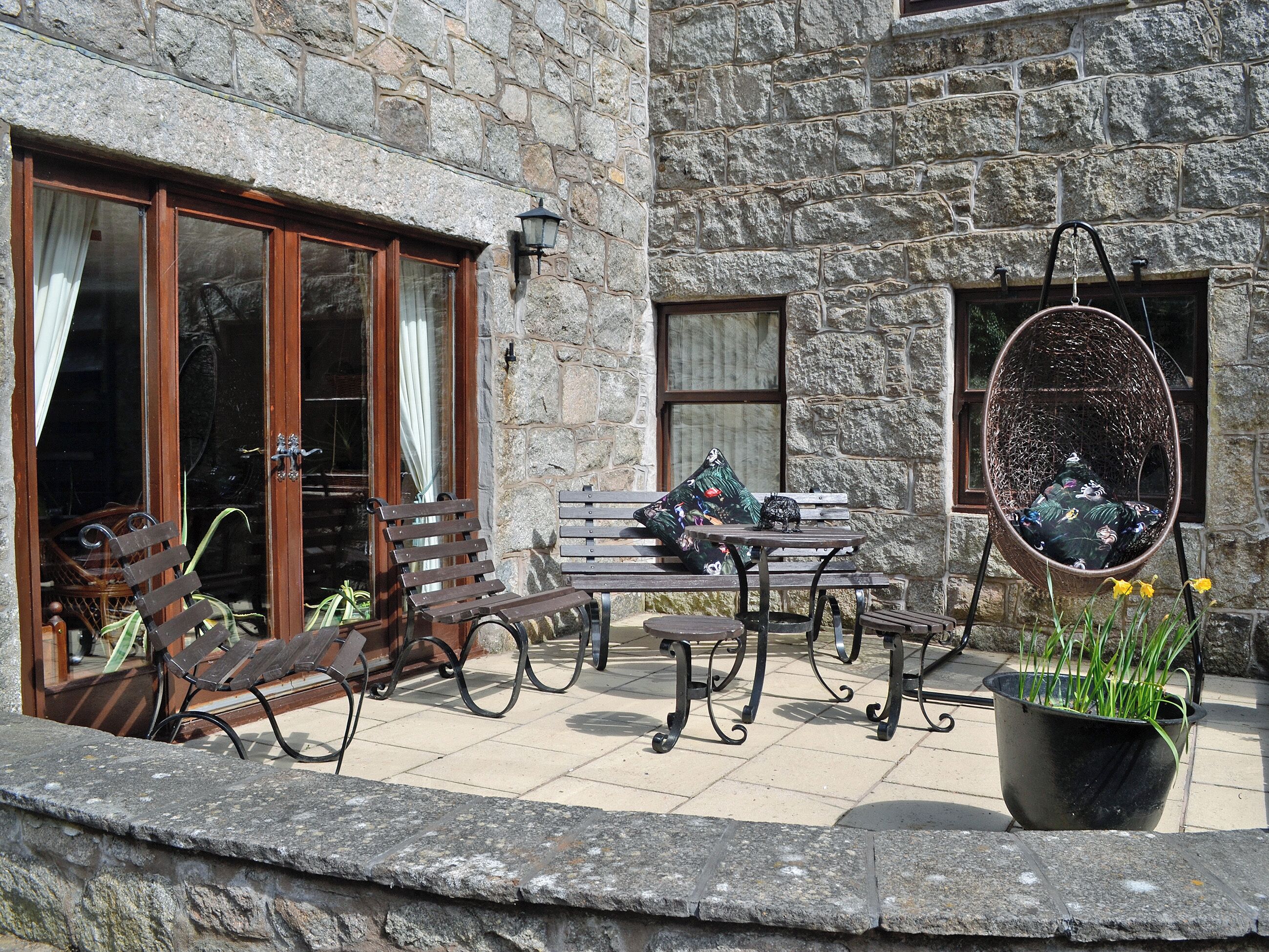 A stone building with a patio area featuring wooden benches, a table, and a hanging chair.