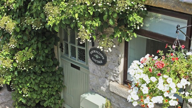 Una cabaña de piedra con puerta verde, un letrero que dice 'Thorncliffe Cottage' y una ventana adornada con flores blancas y rojas.