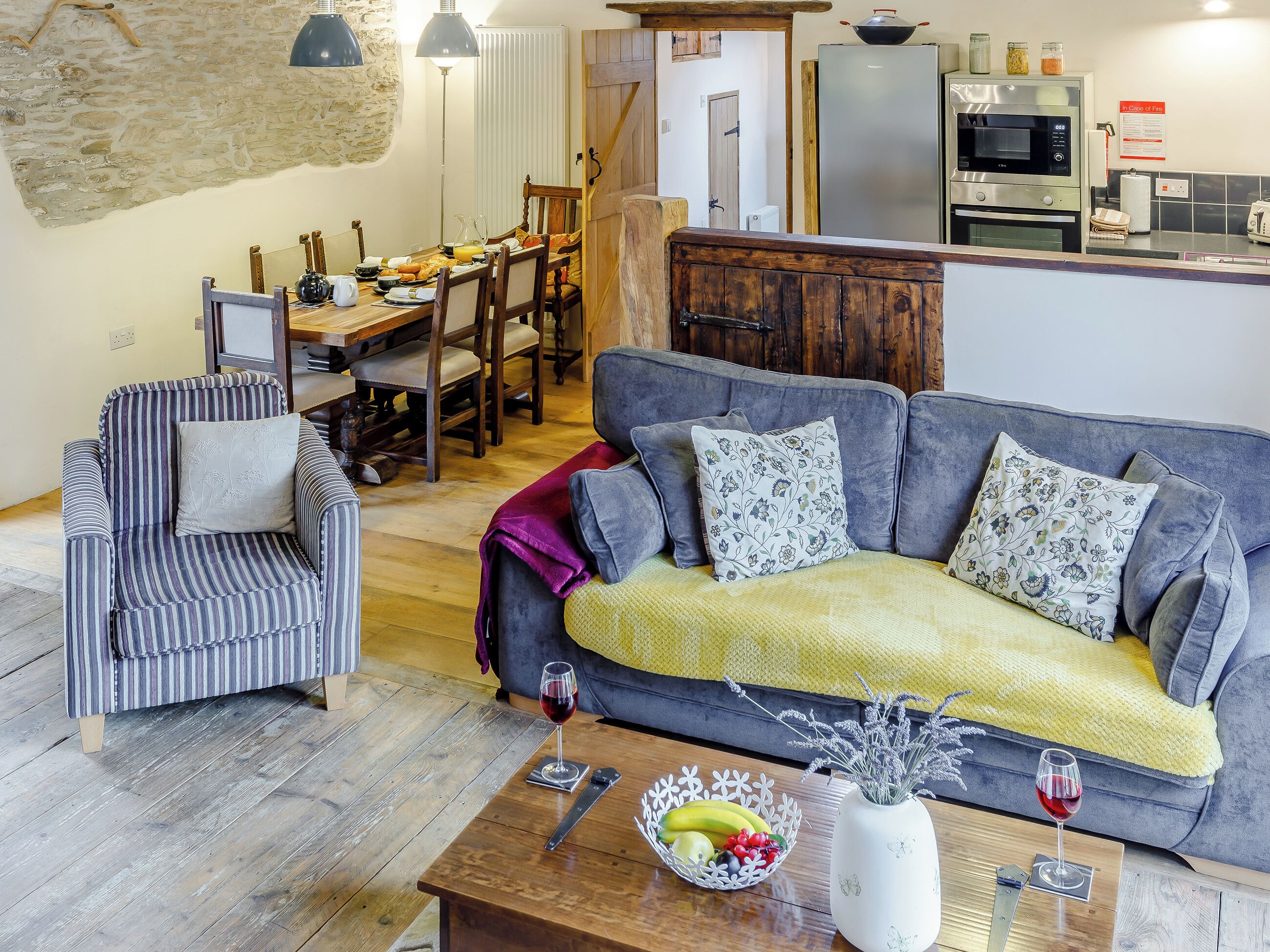 A cozy living room with a sofa, armchair, and dining table. A kitchen with appliances is visible in the background.