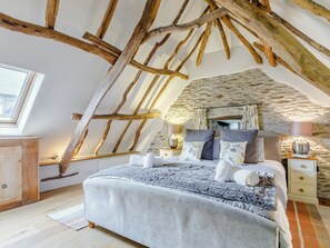 A bedroom with a sloped ceiling, exposed wooden beams, a stone wall, a large bed with pillows and a blanket, bedside tables with lamps, and a window with curtains. - The Threshing Barn - HSSW (Patchole, near Barnstaple)
