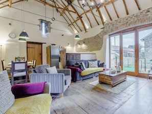 Un vaste espace de vie doté d’un plafond haut, de poutres en bois et d’un mur en pierre. On y trouve un coin repas avec une table et des chaises, un canapé et une table basse sur laquelle est posé un verre à vin.