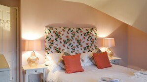 Ein ordentlich bezogenes Bett mit weißen Leinen und zwei orangefarbenen Kissen, ein Nachttisch mit Lampe sowie ein Kopfteil mit floralem Muster.