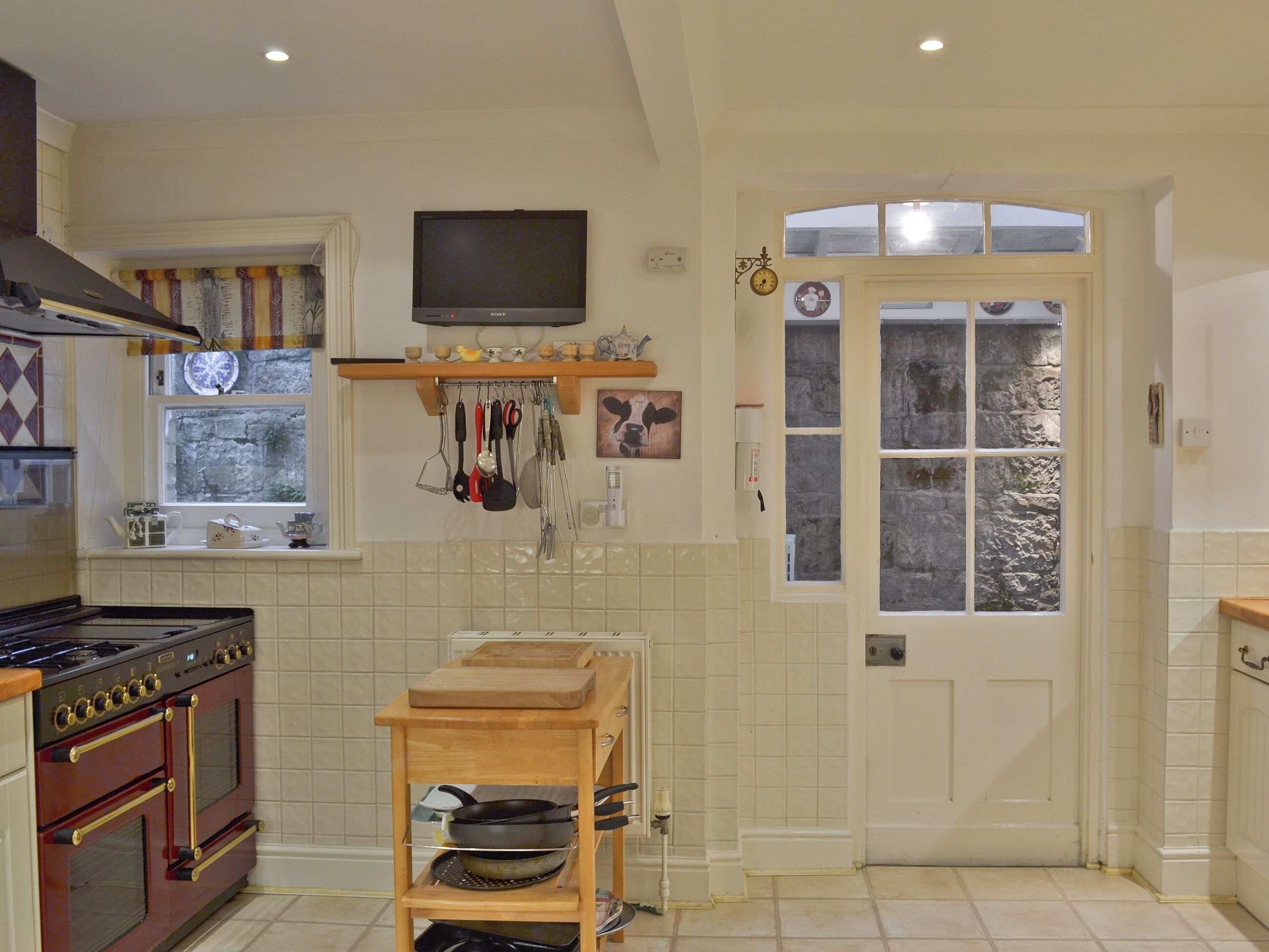 Una cocina con estufa, horno, repisa de madera con utensilios de cocina, un televisor y una ventana con cortinas.