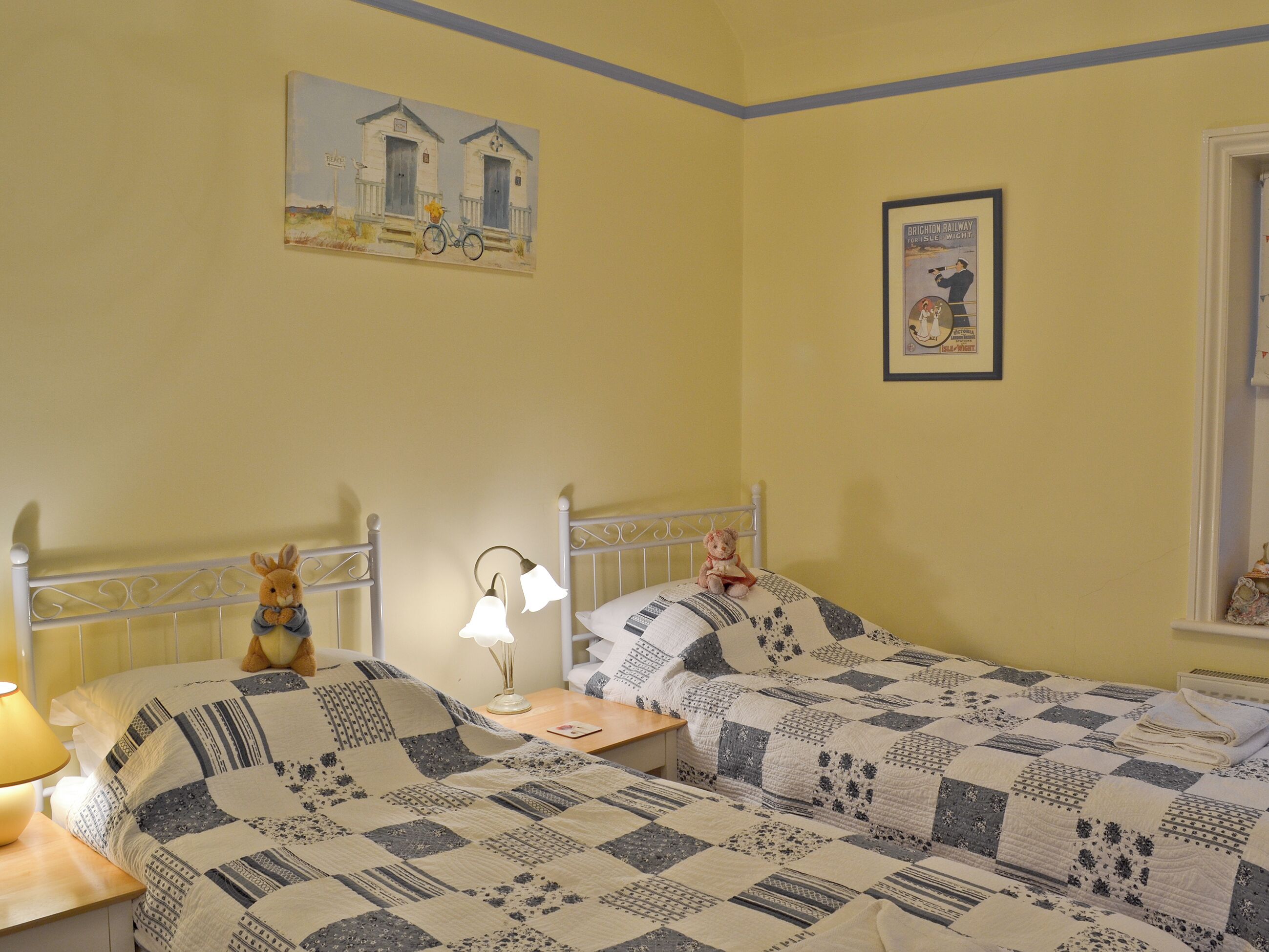 A room with two beds, a nightstand with a lamp, a teddy bear, and two wall art pieces.