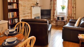 A living room with a stone wall, a bookshelf, a television, a sofa, and a dining area with a table set for two. - 1 bedroom accommodation in Curbar, near Bakewell (Curbar, near Bakewell)