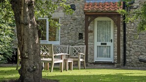 A stone building with a tiled roof, a door with a window, and a small window with a sign. There are wooden chairs and a table outside. - 3 bedroom accommodation in Pickering (Pickering)