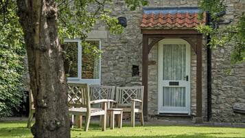 A stone building with a tiled roof, a door with a window, and a small window with a sign. There are wooden chairs and a table outside.