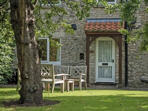 A stone building with a tiled roof, a door with a window, and a small window with a sign. There are wooden chairs and a table outside. - 3 bedroom accommodation in Pickering (Pickering)