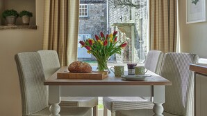 A dining area with wicker chairs, a wooden table, a loaf of bread, a vase with tulips, and a window with a view of a stone building. - 3 bedroom accommodation in Pickering (Pickering)