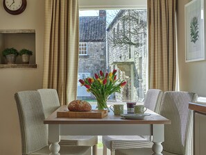 A dining area with wicker chairs, a wooden table, a loaf of bread, a vase with tulips, and a window with a view of a stone building. - 3 bedroom accommodation in Pickering (Pickering)