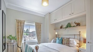 Ein Schlafzimmer mit Bett, Nachttisch, Vorhängen am Fenster, Heizkörper, Ablagefläche mit Dekoration und einer Pflanze.