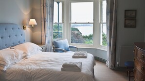 A bedroom with a large bed, a chair, a dresser, and a view of a body of water through a bay window. - 8 bedroom accommodation in Porthpean, near Charlestown (Porthpean, near Charlestown)