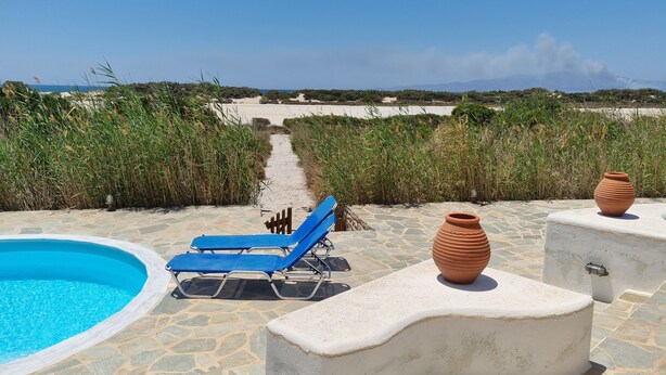 Un área de piscina con sillas de descanso azules y macetas de terracota, rodeada de baldosas de piedra y un sendero hacia la playa.