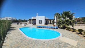 Una piscina con sillas reclinables azules, un patio de piedra y un edificio blanco de fachada pétrea.