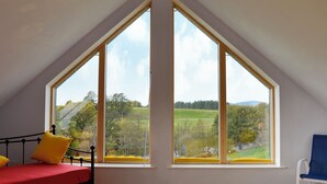 A room with a sloped ceiling, a bed with yellow pillows, a chair, and large windows offering a view of a green landscape.