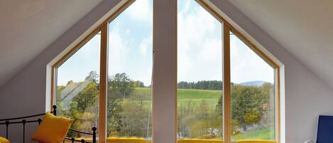 Una habitación con techo inclinado, una cama con almohadas amarillas, una silla y ventanas amplias que ofrecen vistas a un paisaje verde.