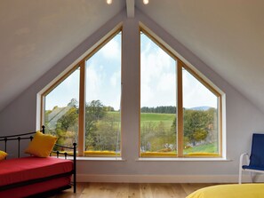 Une pièce avec un plafond en pente, un lit avec des coussins jaunes, une chaise et de grandes fenêtres donnant sur un paysage verdoyant.