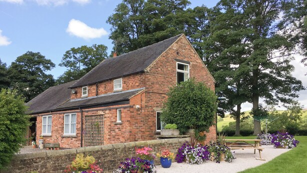 Une maison en briques avec un toit en pente, entourée d’un chemin de gravier et de parterres de fleurs aux couleurs vives.