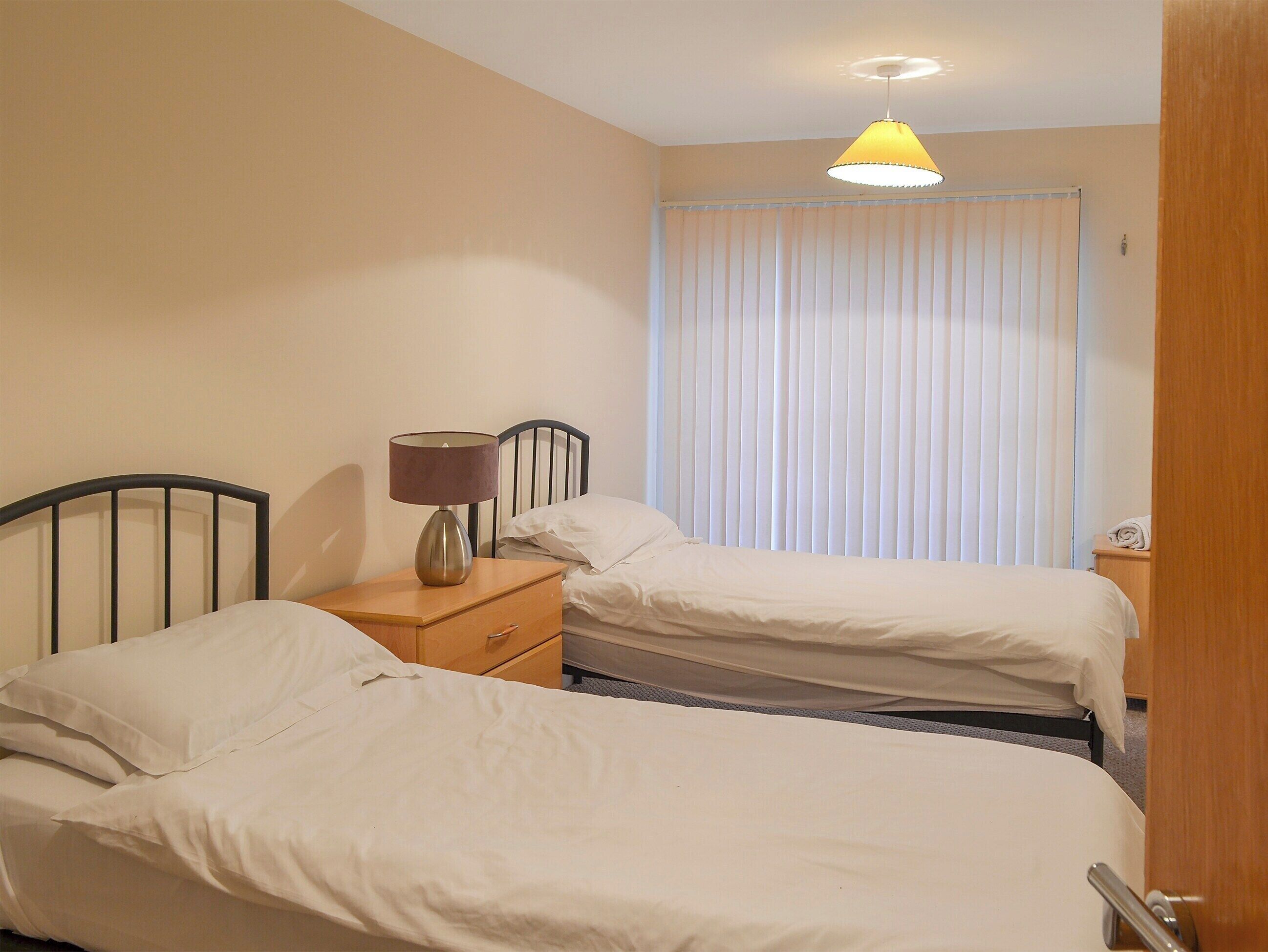 A hotel room with two single beds, a bedside table with a lamp, a window with blinds, and a ceiling light.