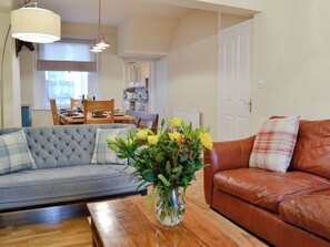 A living room with a sofa, a coffee table, a vase with flowers, a dining area with a table and chairs, and a kitchen in the background.