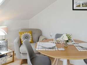 A dining area with a round table set for two, a sofa with decorative pillows, a side table with a lamp, and a framed picture on the wall.